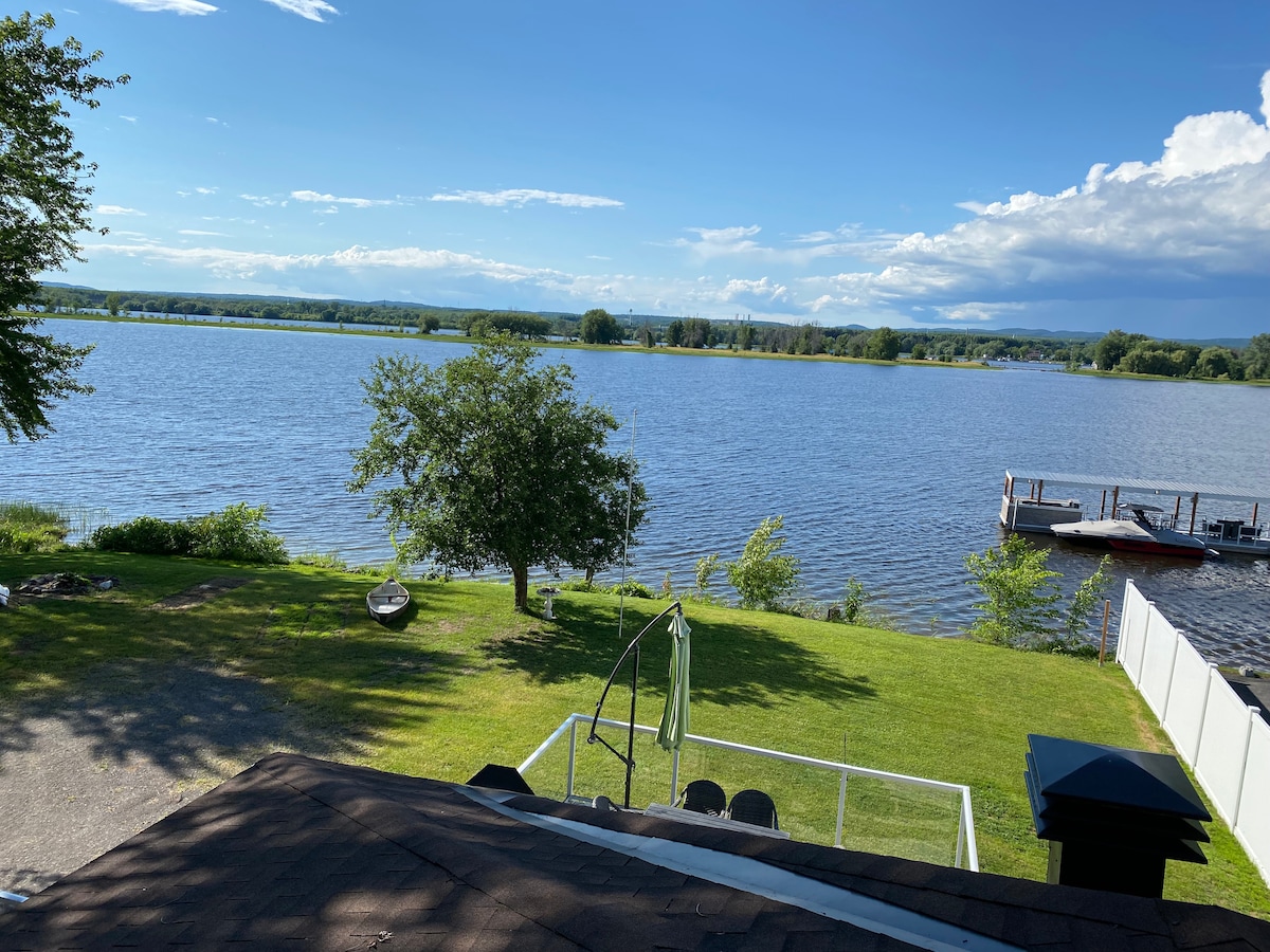 A serene view of the Ottawa River is captured from a raised vantage point. Lush greenery surrounds the water, while a small dock and boat are visible in the distance. Sunlight reflects off the rippling surface, creating a calm and inviting waterway atmosphere.