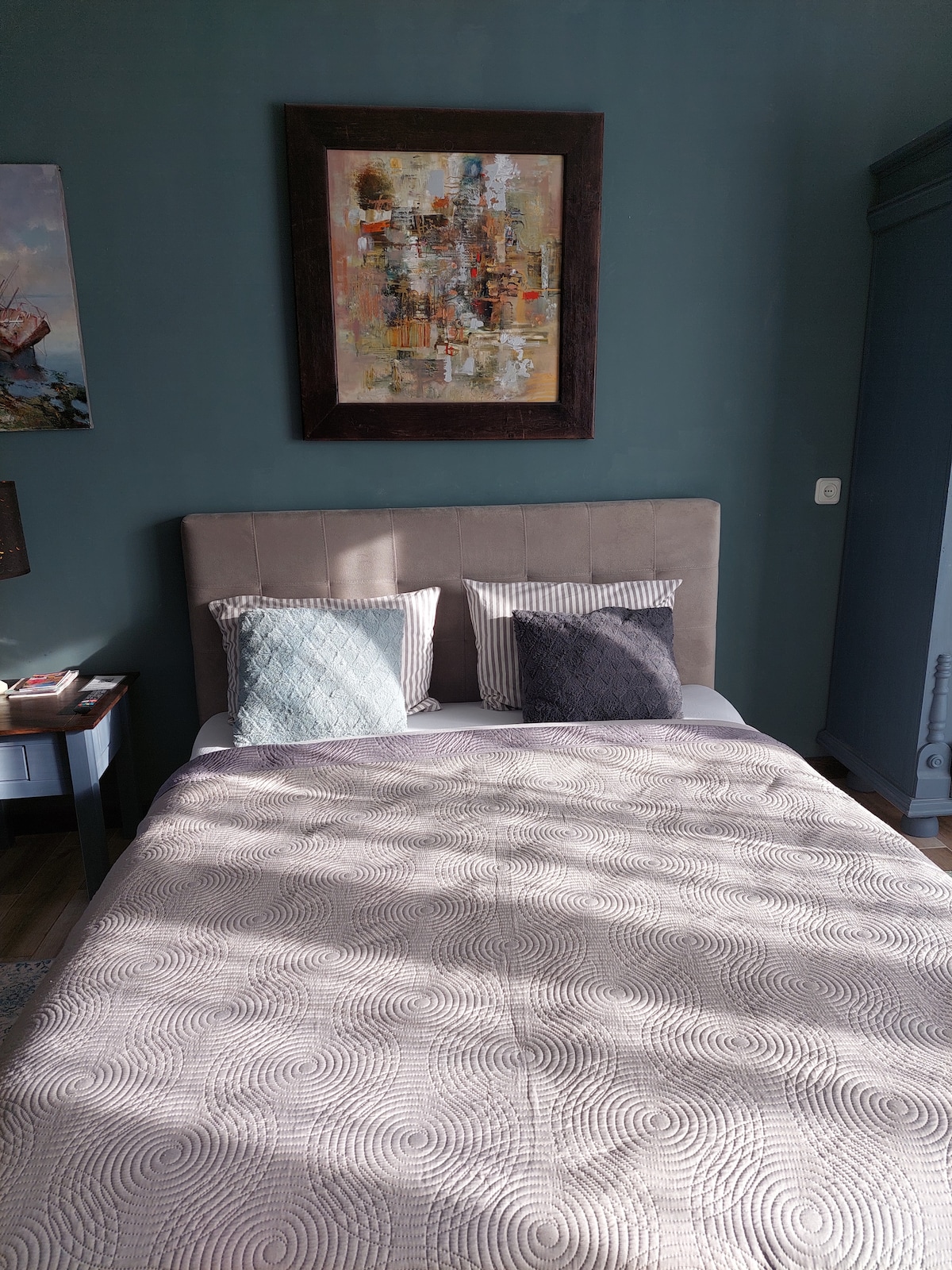 A serene sleeping area features a spacious bed adorned with a textured blanket and various decorative pillows. A piece of abstract artwork hangs above the headboard, complementing the muted green walls. Natural light filters softly, enhancing the calm ambiance.