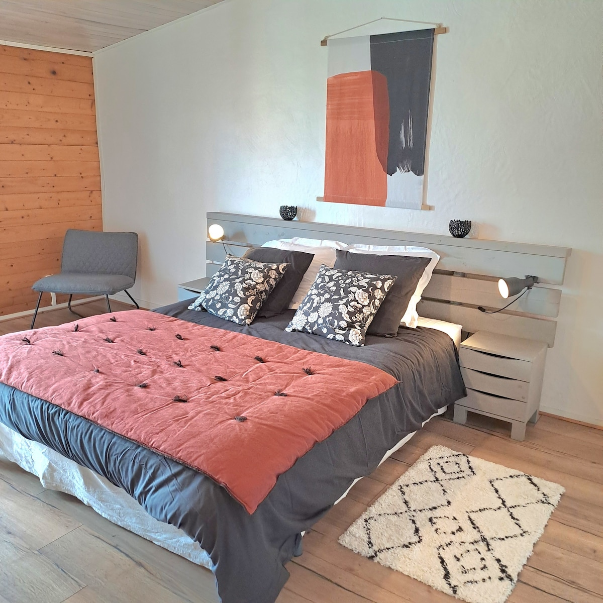 A spacious sleeping area features a double bed adorned with a textured coral blanket and decorative pillows. A minimalist artwork is displayed above the bed, with a cozy chair positioned nearby, complemented by natural wood accents and warm wooden flooring.