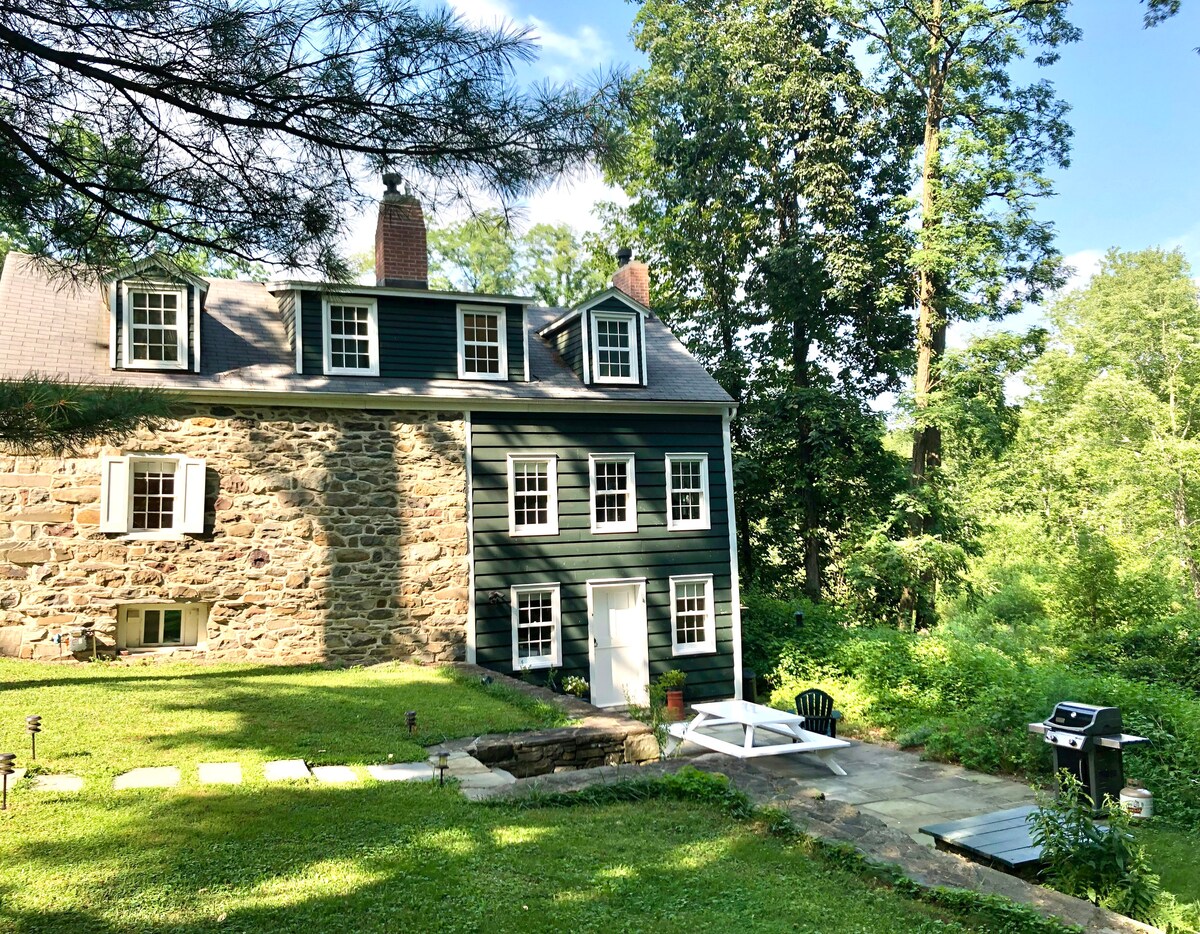 The exterior of the historic stone house is highlighted by its three floors, featuring stone and clapboard siding. A private terrace with a picnic table and propane grill is visible on the patio, surrounded by lush greenery and trees.