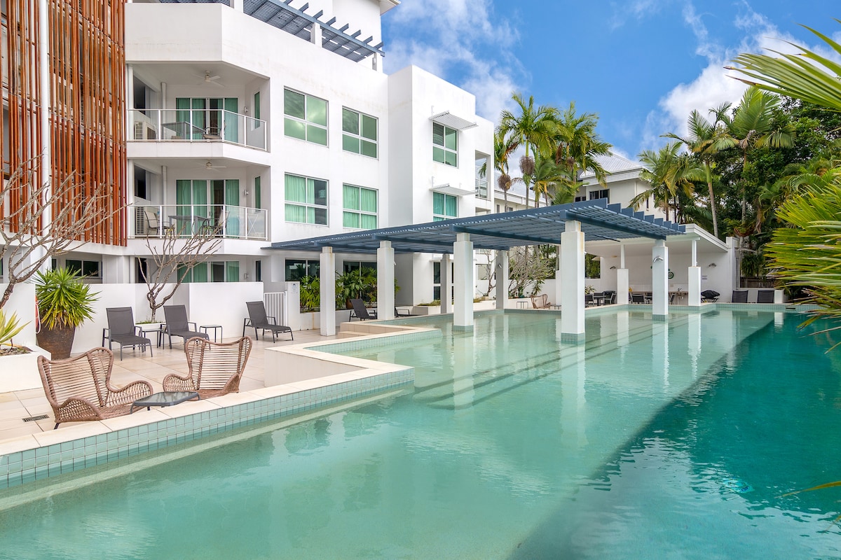 A large swimming pool with a lap lane is surrounded by tropical landscaping and modern architecture. Comfortable seating areas are available under a shaded structure, enhancing the inviting ambiance of the outdoor space.