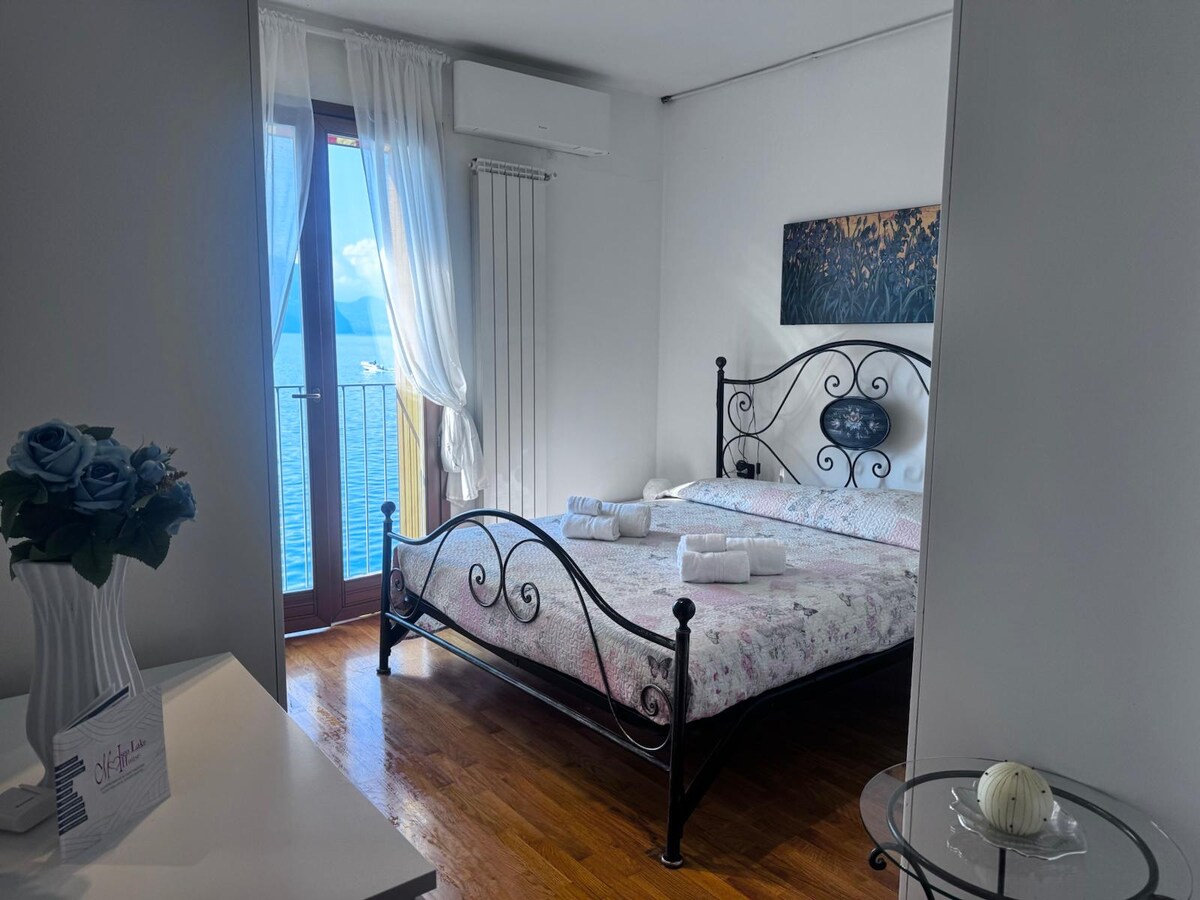 A well-lit bedroom features a decorative iron bed with floral bedding. Large glass doors open to a balcony offering lake views, while light curtains gently frame the entrance. Wooden flooring complements a subtle color palette, and a painting adorns the wall above the bed.