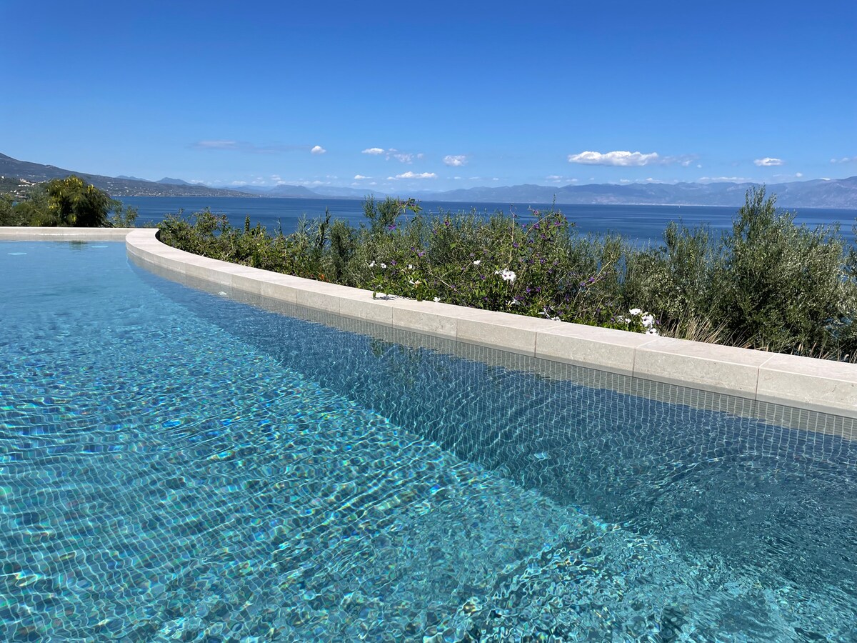 A serene salt-water pool reflects the clear blue sky and the expansive Messinian bay. The pool's edge appears to blend seamlessly with the horizon, surrounded by lush greenery and flowering plants that enhance the natural setting.