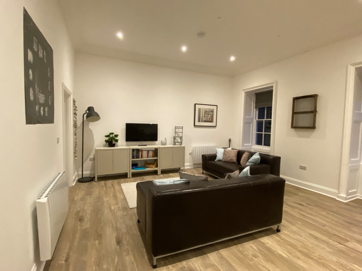 A spacious living area is showcased, featuring a comfortable brown leather sofa and a light-colored entertainment unit. A television is mounted on the wall opposite. Natural light floods in from the large windows, illuminating the minimalistic decor and warm wood flooring.