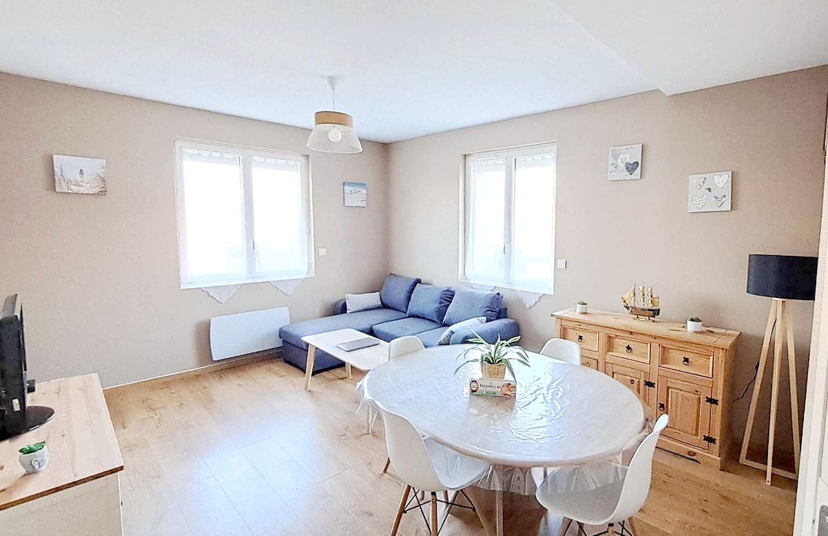 A light-filled living area features a comfortable navy blue sofa, a round dining table surrounded by white chairs, and a wooden sideboard. Natural light flows through large windows, highlighting the warm-toned walls and creating a welcoming space for relaxation and dining.