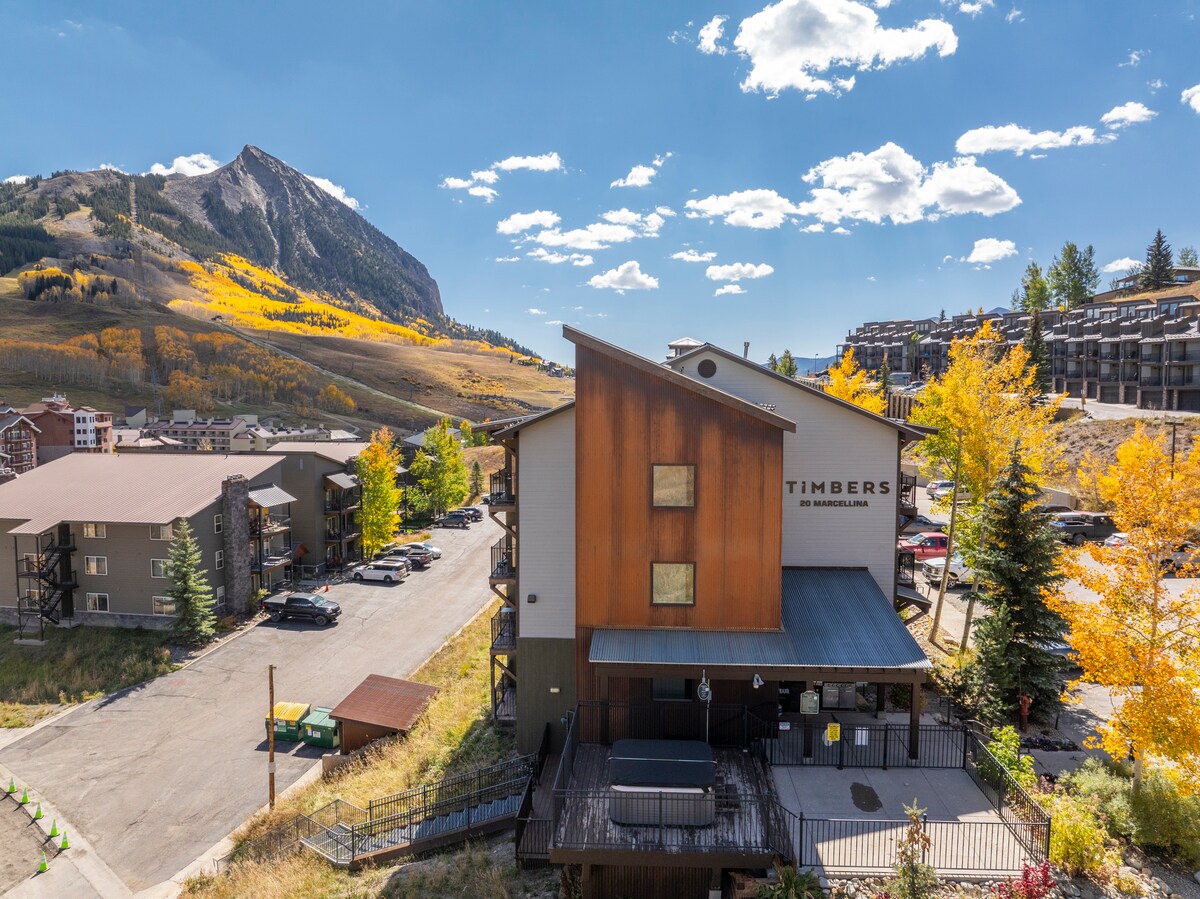 An aerial view showcases the condo building nestled among trees, with vibrant fall foliage in the background. The spacious deck features a hot tub, and neighboring buildings frame the scene. The majestic mountain peak rises in the distance under a clear blue sky.