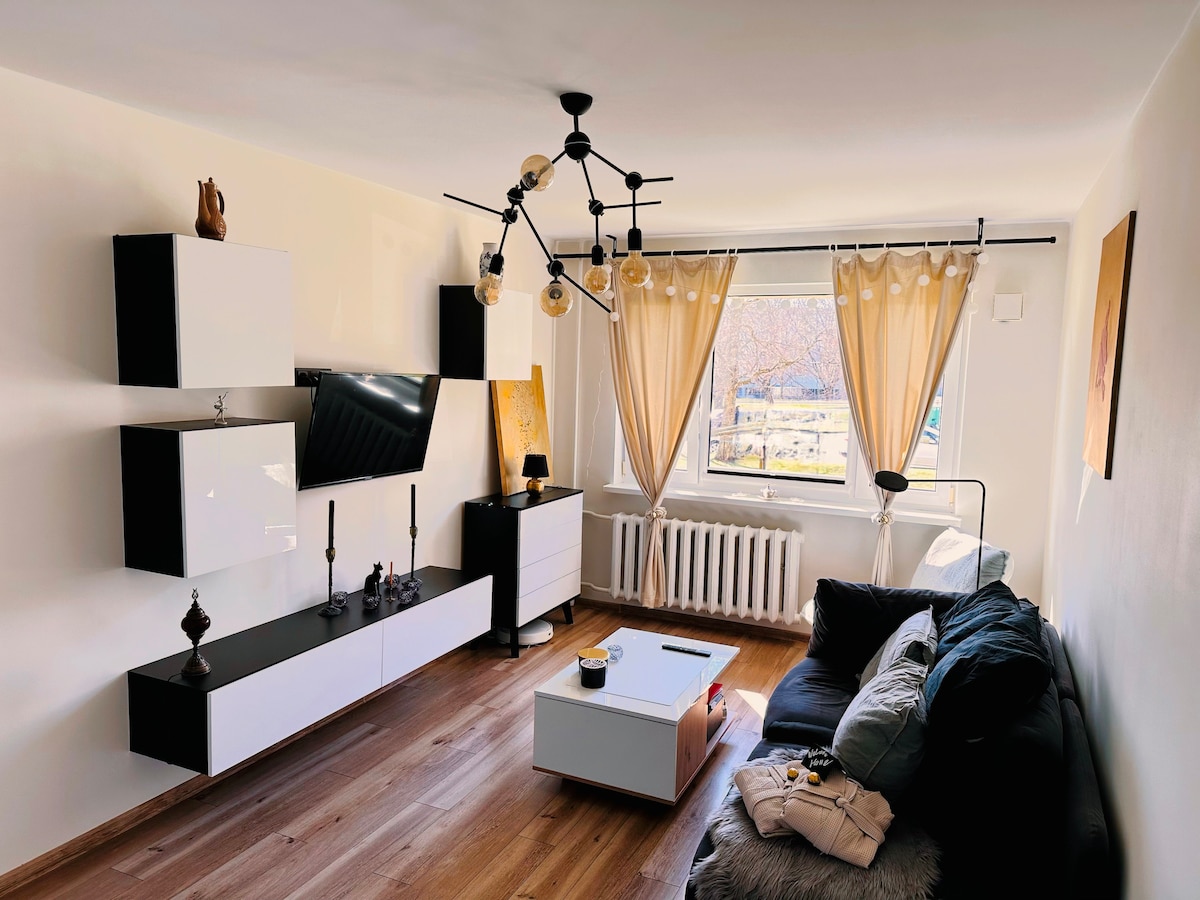A bright living space features a comfortable dark sofa and a sleek white coffee table, complemented by decorative items. A wall-mounted television is positioned above a low white cabinet. Natural light enters through large windows adorned with light curtains, providing a view of the outdoor area.