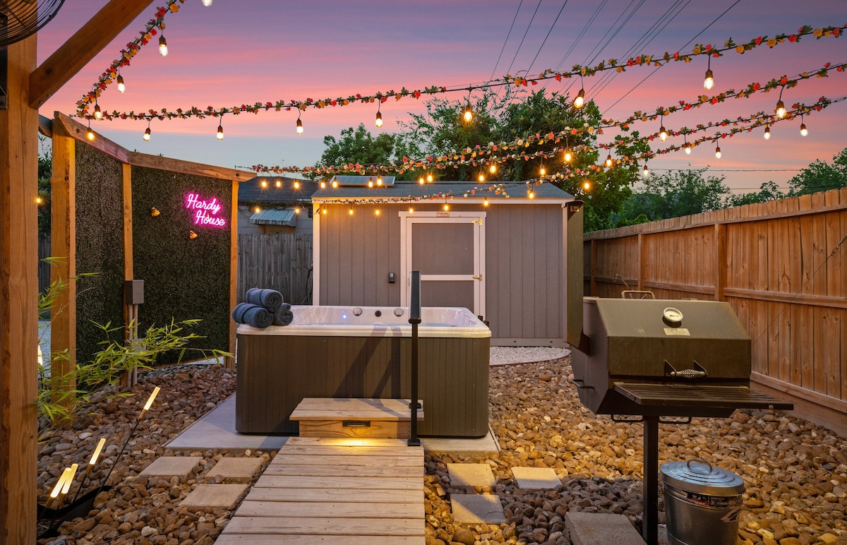 A serene outdoor space features a hot tub beneath twinkling string lights, providing a cozy ambiance. A grill and wooden walkway are visible, creating a perfect setting for relaxation and entertainment. The space is surrounded by a wooden fence, offering privacy.