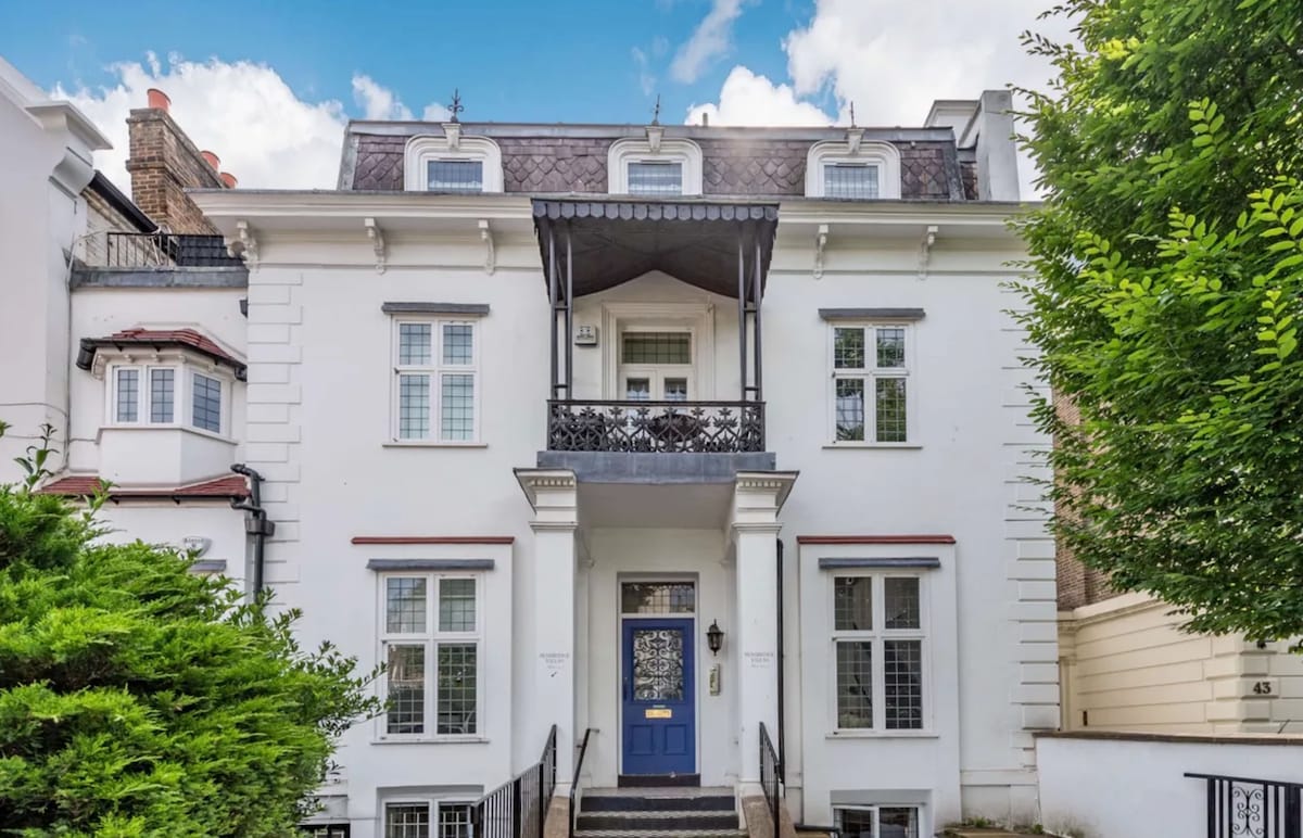 A stately white building with a classic architectural design is highlighted, featuring large windows and a decorative balcony. The entrance is flanked by greenery, with steps leading to the inviting front door. The roof includes charming gables, adding character to the structure.