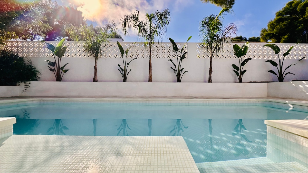 The private swimming pool is framed by green plants and a white stone wall. The clear blue water reflects the sunlight, creating a serene atmosphere. The pool area offers a retreat for relaxation with ample space for lounging.