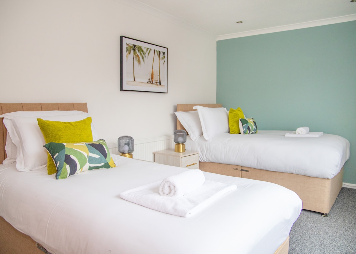 The bright bedroom features two spacious beds topped with white linens and decorative pillows in green and black. A bedside table holds a lamp, and a framed tropical artwork adds a touch of colour to the wall. Natural light fills the room from a window.