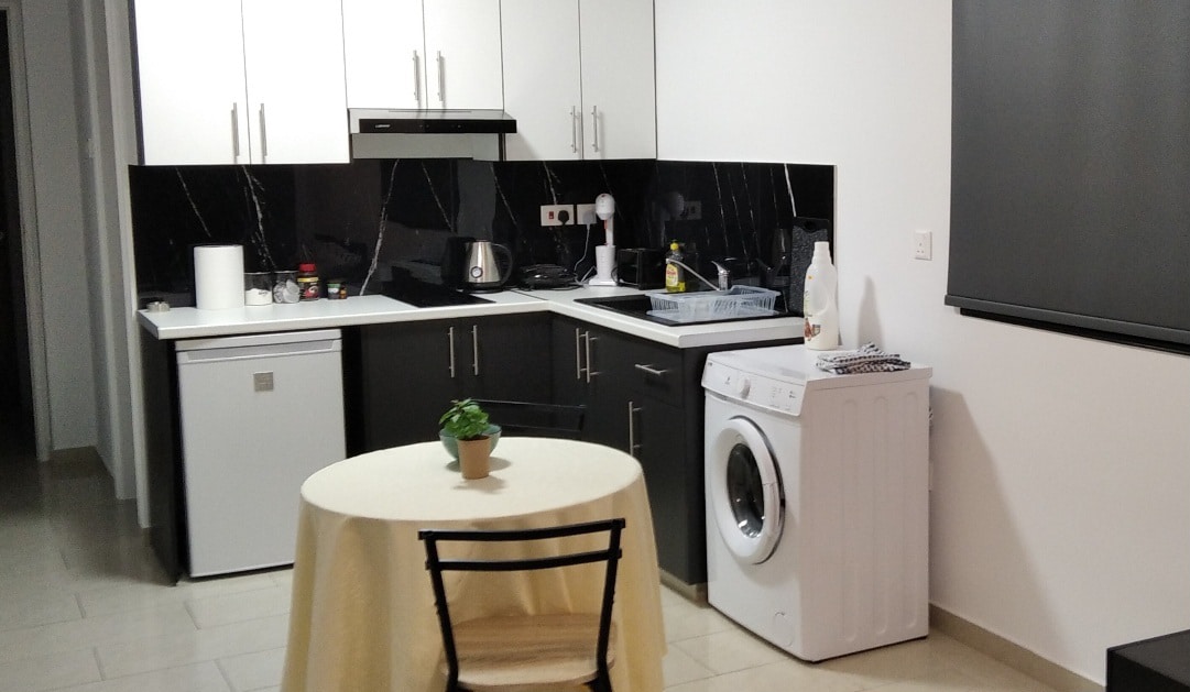 A modern kitchen area is equipped with sleek black and white cabinetry, featuring a refrigerator, stove, and sink. A round dining table with a light-colored cloth is paired with a black chair. A washing machine is positioned against the wall, and a small plant adds a touch of greenery.