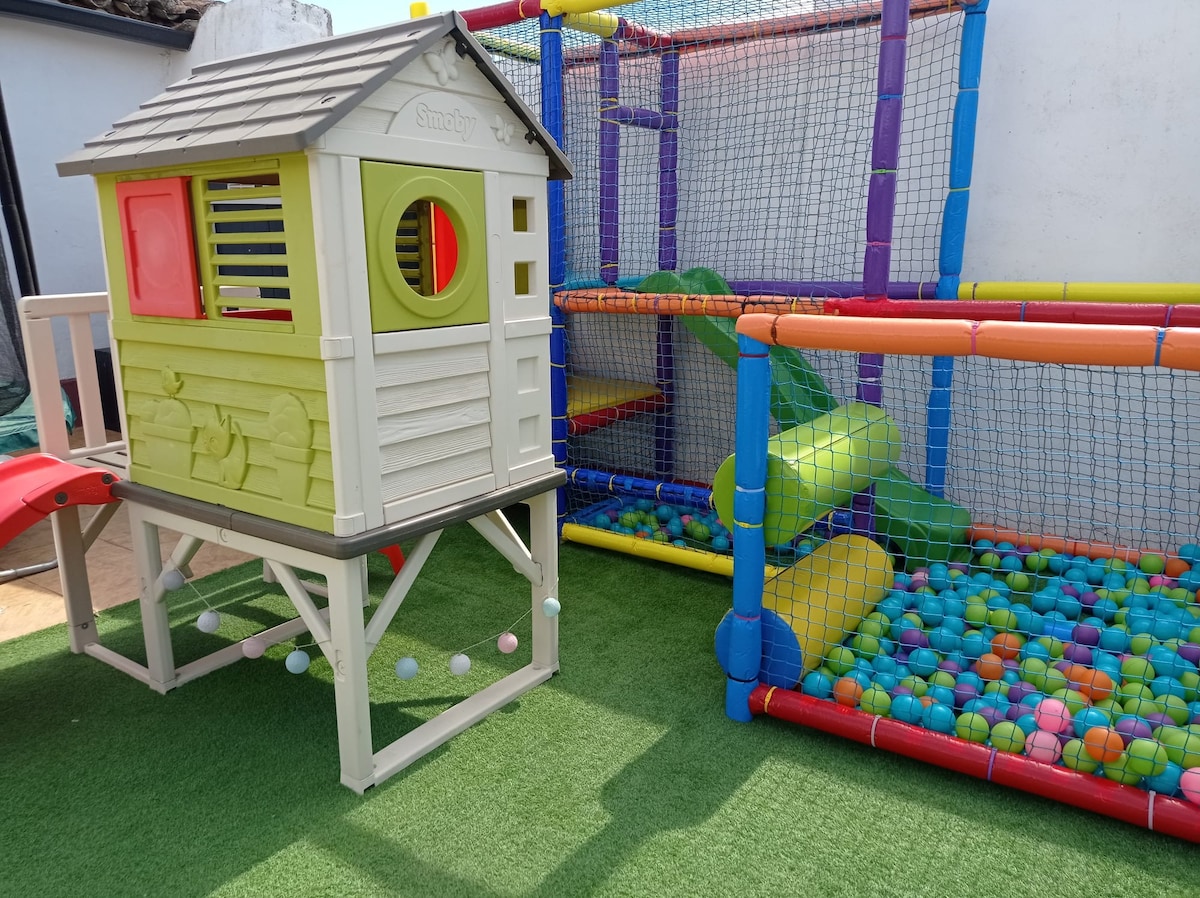 A children's play area features a colorful plastic playhouse with a slide alongside a vibrant ball pit filled with assorted colorful balls. An adjacent climbing structure provides additional play opportunities, all set on a soft artificial grass surface.