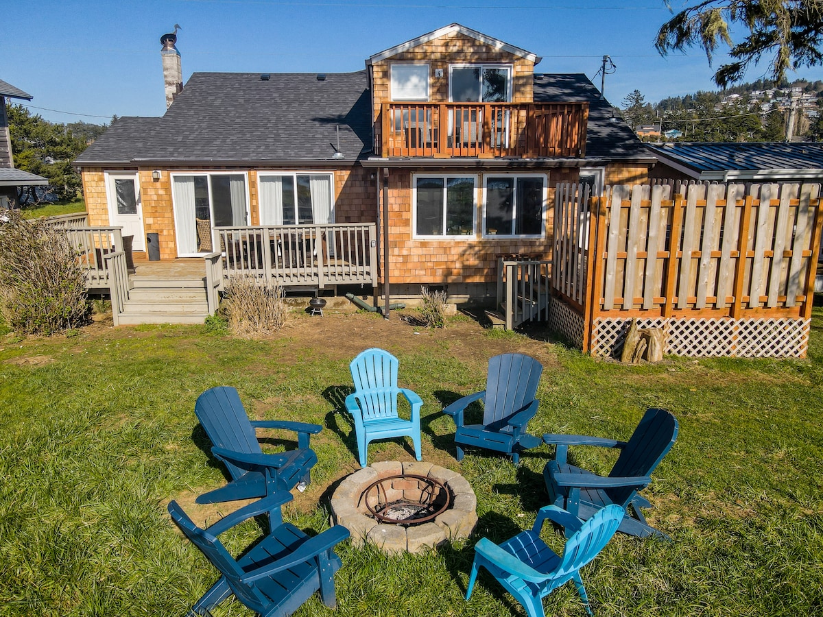 The spacious backyard features a circular arrangement of eight colorful adirondack chairs surrounding a stone fire pit. The multi-level house is visible in the background, showcasing a wooden deck and large windows that invite sunlight into the living space.