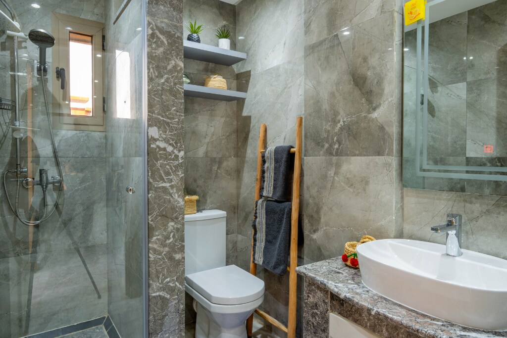 A modern bathroom features a glass shower with a rainfall showerhead, surrounded by sleek marble tiles. A white sink is positioned on a stone countertop, while a toilet is located nearby. Wooden shelves display decorative plants, and towels are neatly arranged on a minimalist ladder.