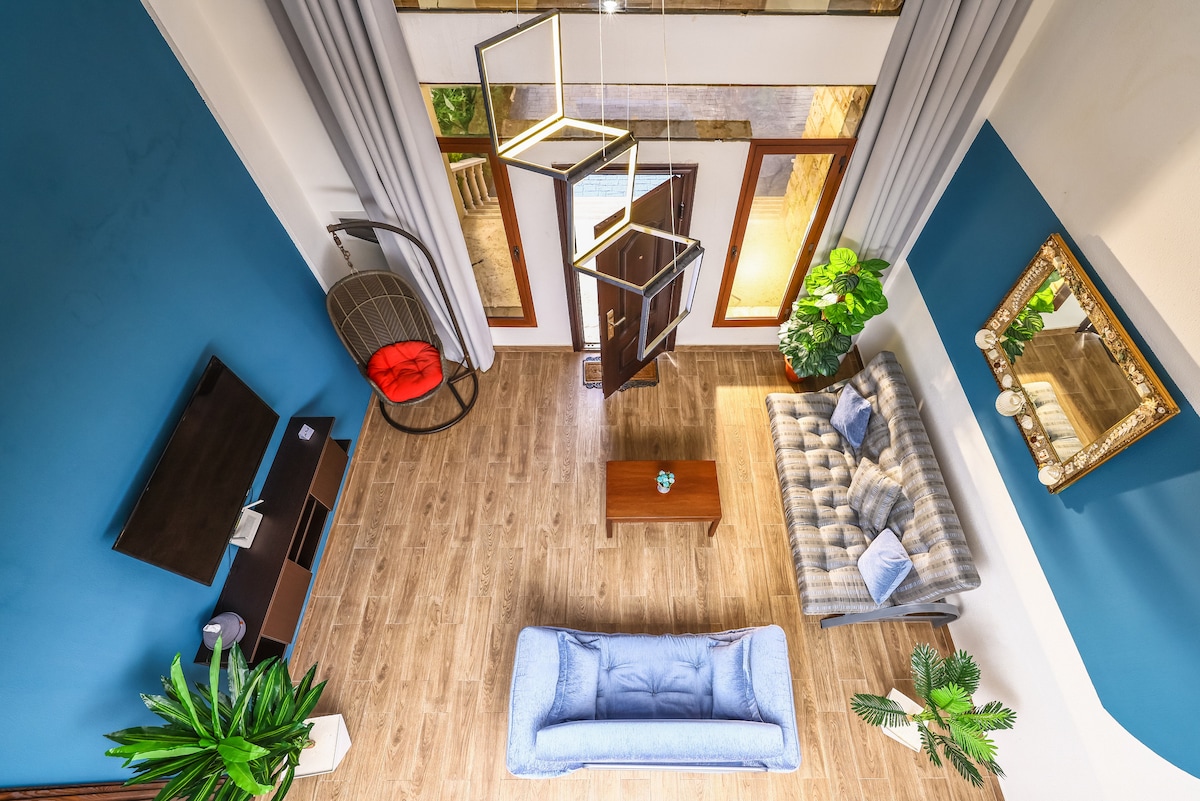 A spacious living area is presented with a blend of natural wood and vibrant blue walls. A comfortable sofa and coffee table are positioned centrally, surrounded by decorative plants. Large glass doors allow ample natural light to fill the space, enhancing the inviting atmosphere.