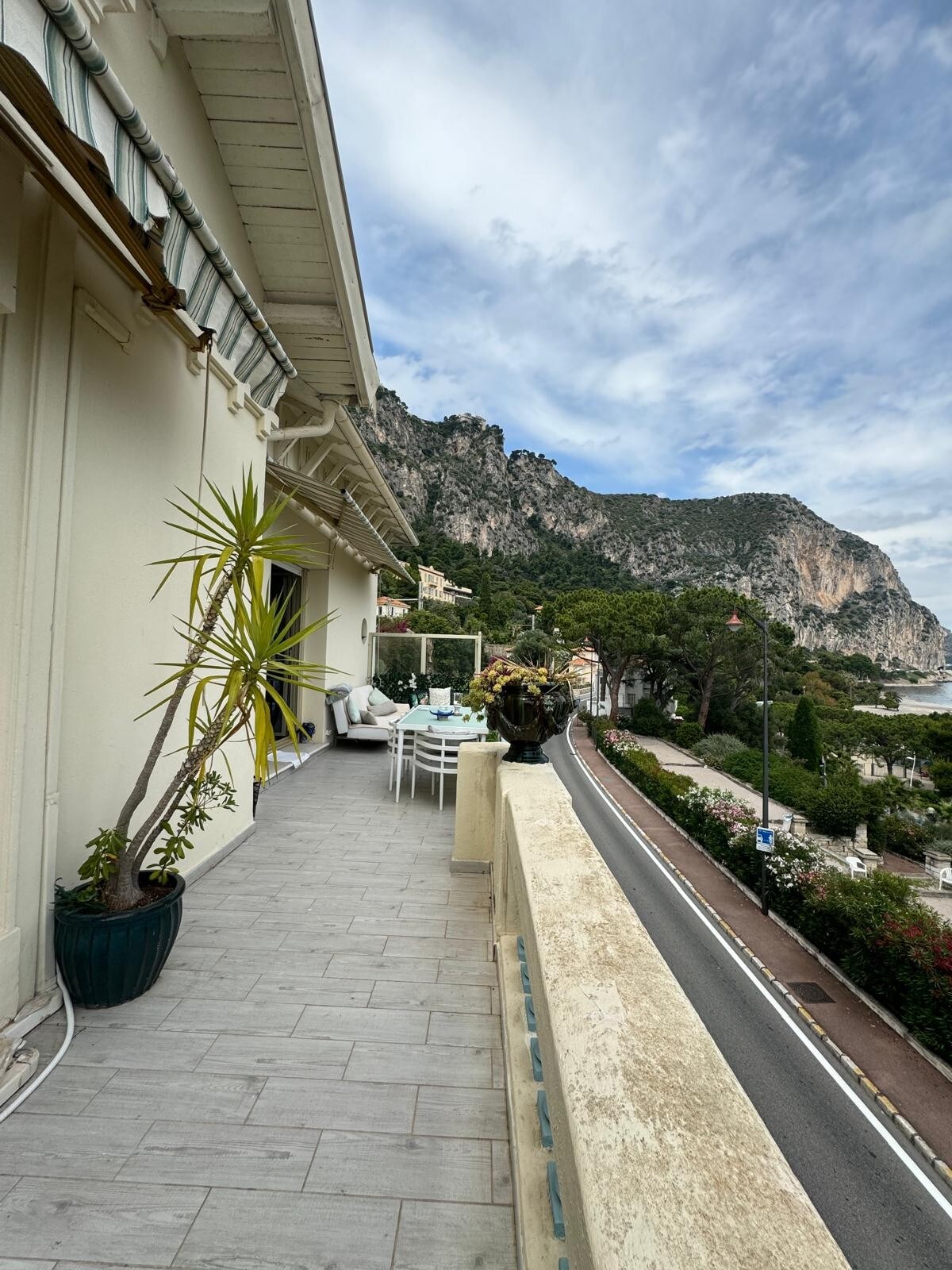 A spacious terrace is visible, featuring a dining table surrounded by several chairs. Potted plants and decorative flowers enhance the area. In the background, a rugged mountain landscape is seen, adding a natural backdrop to the outdoor space.