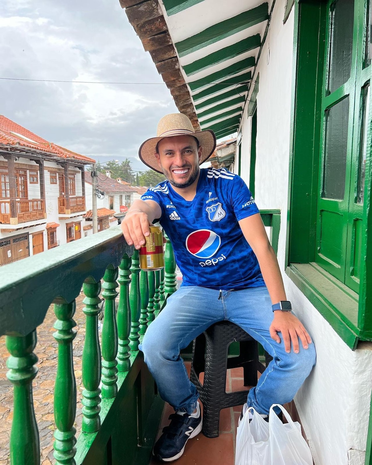 A person in a blue sports jersey and a straw hat is seated on a balcony chair, overlooking a cobbled street. The green railing frames the view, while a shopping bag is positioned on the ground. The scene captures a relaxed moment in an inviting outdoor space.