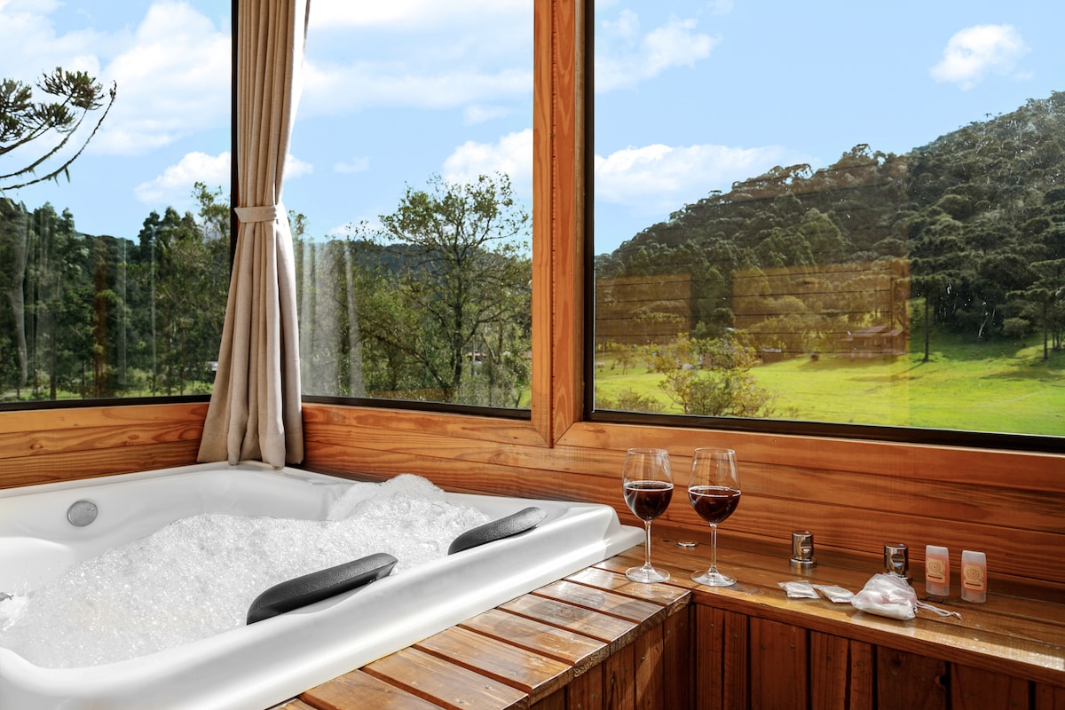 A spacious hot tub is situated near large windows, offering views of the surrounding greenery. Wooden accents complement the serene setting, and two glasses of red wine rest on a wooden ledge, alongside various amenities for relaxation.