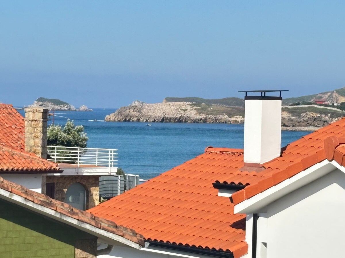 A coastal view captures the serene blue sea meeting rocky formations in the distance. Roofs with orange tiles frame the scene, while a white chimney stands prominently among the houses, indicating a tranquil seaside atmosphere.
