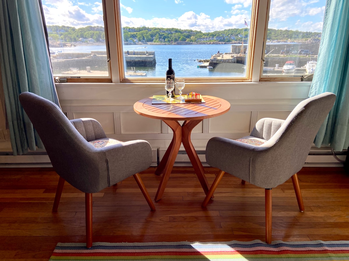 A cozy cafe table is positioned between two modern chairs, overlooking the inner harbor. Natural light floods the space through large windows, creating a warm ambiance. A vibrant area rug adds a touch of color, complementing the serene view of the water and boats.
