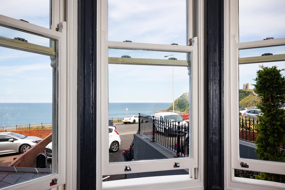 A set of double windows frames a panoramic view of the sea, with cars parked in a landscaped area below. The blue sky with wispy clouds contrasts against the shoreline, enhancing the scenery. Gentle greenery is visible to the right, completing the coastal view.