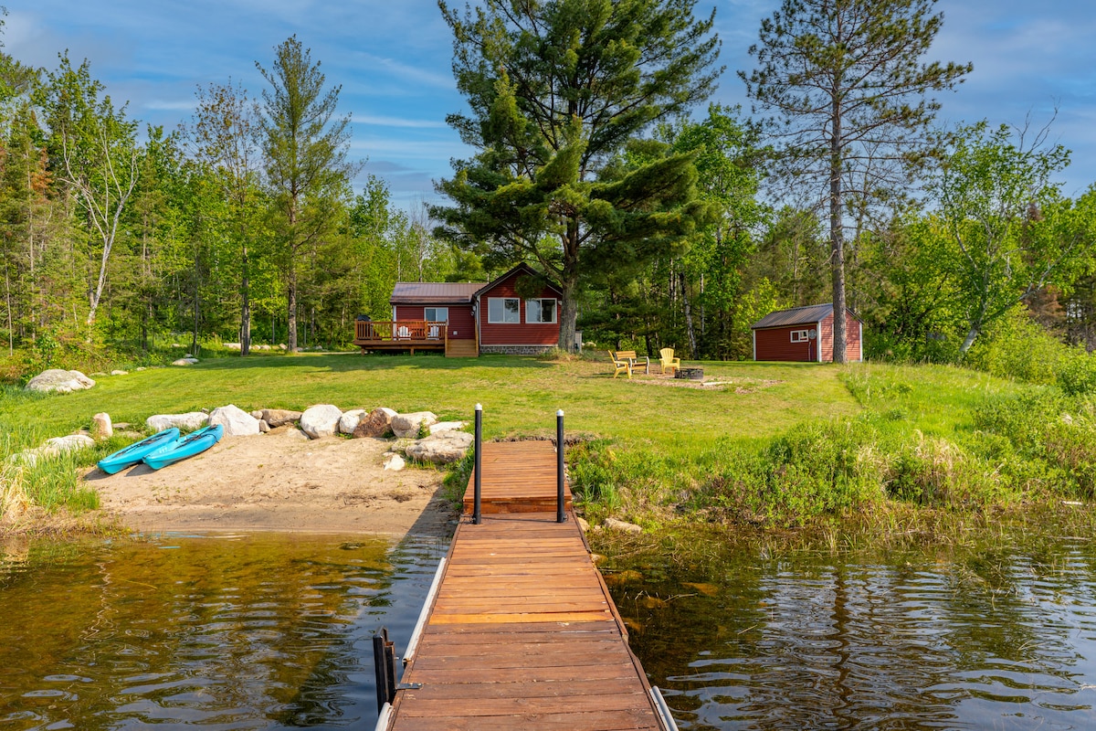 A private dock extends into the calm waters of Burntside Lake, leading to a grassy area featuring a cozy cabin and a small shed. The surrounding landscape includes trees and a sandy beach area, with two kayaks resting on the shore.