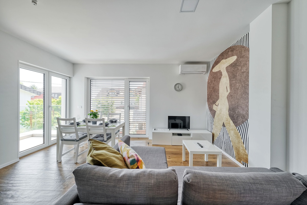 A spacious living room features a comfortable sofa with colorful pillows, complemented by a modern dining area with a table and chairs. Natural light fills the space through large glass doors, providing views of the balcony. A stylish wall art piece adds a unique touch.
