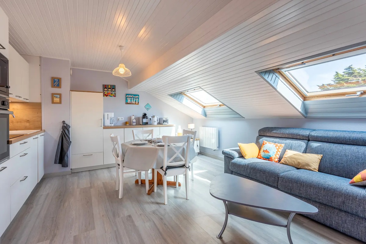 A bright living area features a spacious gray couch adorned with colorful cushions. A round coffee table sits nearby, while a dining area with white chairs and a light wooden table is visible. Velux windows provide natural light, illuminating the welcoming, neutral tones throughout.