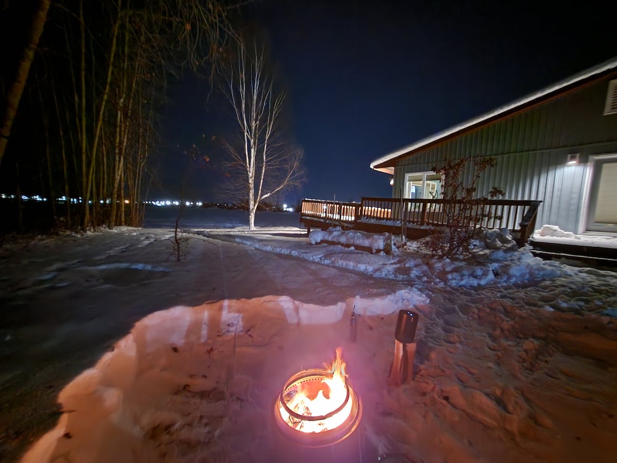 A fire pit is positioned in the foreground, surrounded by smooth, packed snow. The villa's exterior is visible in the background, featuring a wrap-around deck and large windows, with trees lining the property. The night sky is illuminated by distant lights.