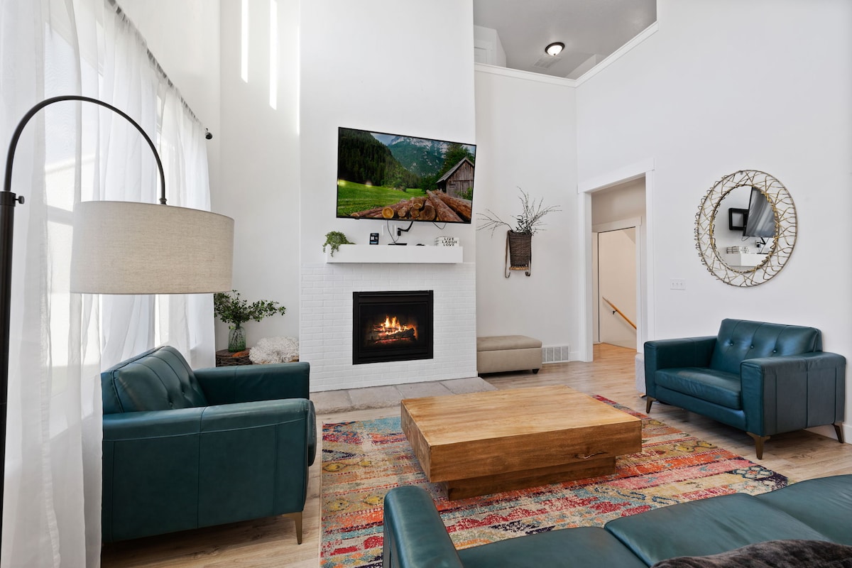 A bright living area features a fireplace beneath a mounted television, framed by light-colored walls. Dark green leather couches face a wooden coffee table placed on a colorful area rug. Natural light filters through sheer curtains, enhancing the spacious feel of the room.