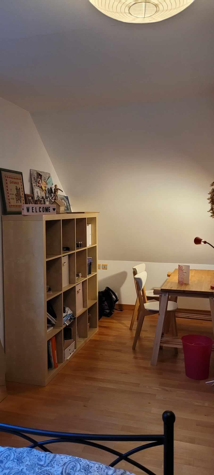 The room features a wooden bookshelf filled with books and decor items, positioned against a sloped wall. A small round table and white chairs are arranged nearby. Soft lighting creates a calm ambiance, and the wooden floor adds warmth to the space.