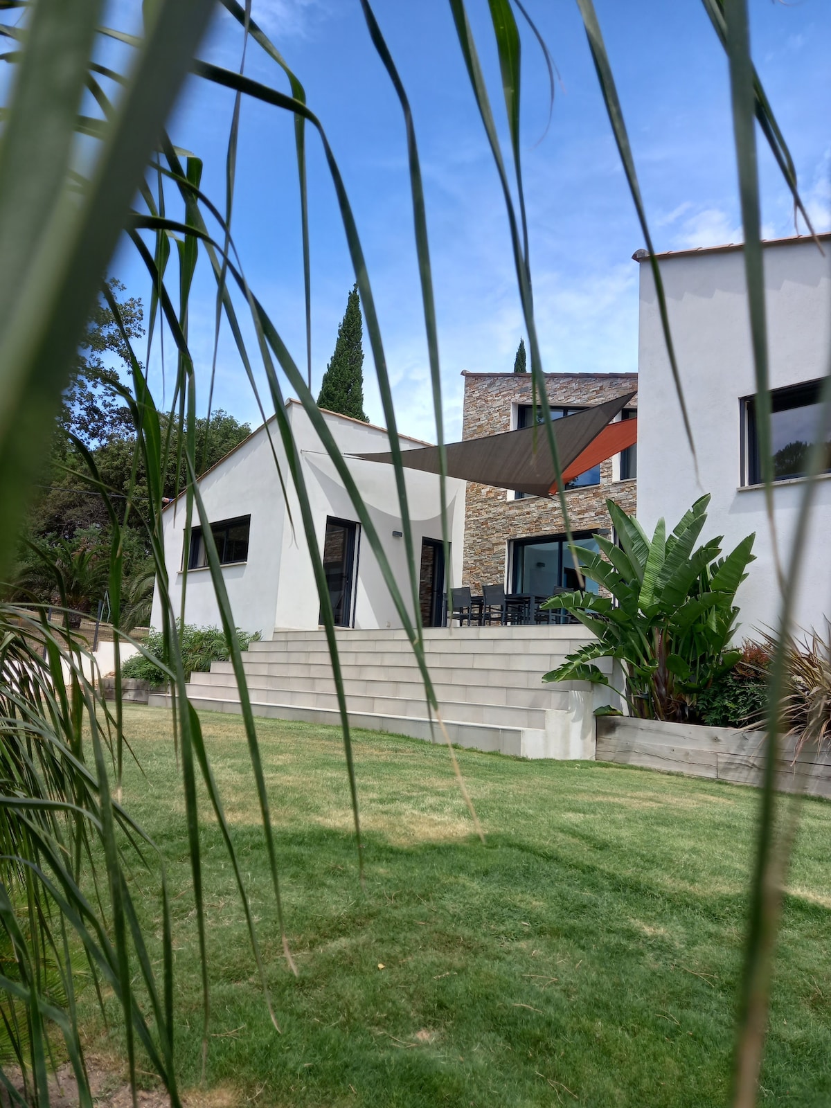 The exterior of a modern villa is framed by lush greenery. The structure features large windows and a shaded outdoor dining area. Steps lead down to the manicured lawn, providing a serene view of the garden surroundings.