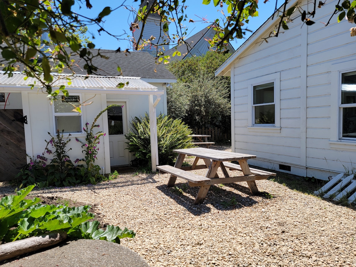 An outdoor area features a wooden picnic table surrounded by gravel and low shrubs. Nearby, a doorway provides access to a small building, while a larger structure is visible in the background, creating a pleasant space for relaxation.
