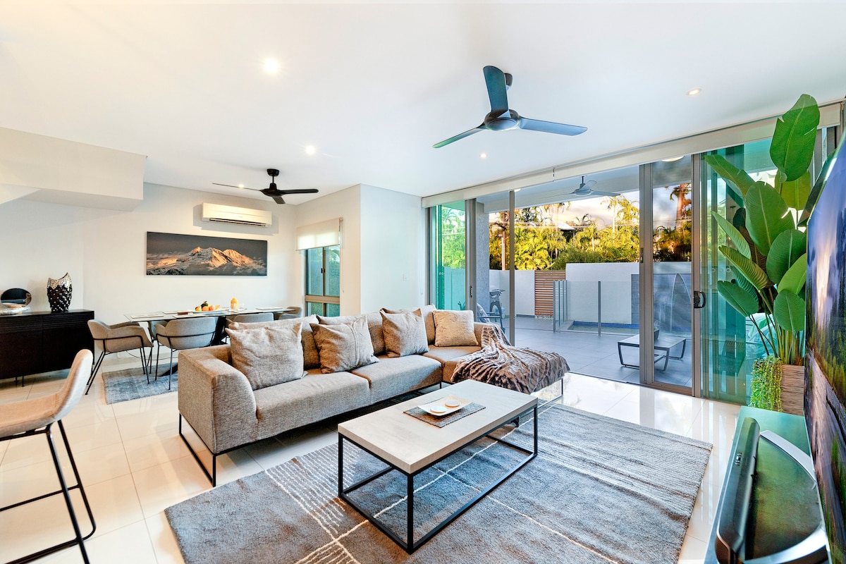 A modern living area features a spacious grey sofa with cushions, complemented by a sleek coffee table. Large glass doors provide an abundance of natural light and open to a private outdoor space, while a ceiling fan adds comfort to the environment.