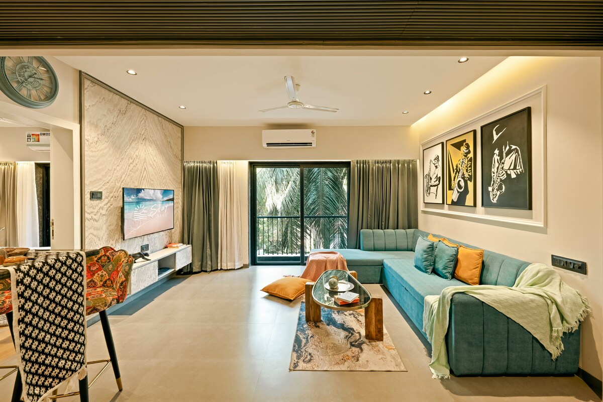 The open-plan living and dining area features a cushioned sofa, a coffee table, and a wall-mounted smart flatscreen TV. Natural light floods in through tall sliding windows, offering views of the surrounding greenery. Colorful art pieces adorn the walls, contributing to a welcoming ambiance.