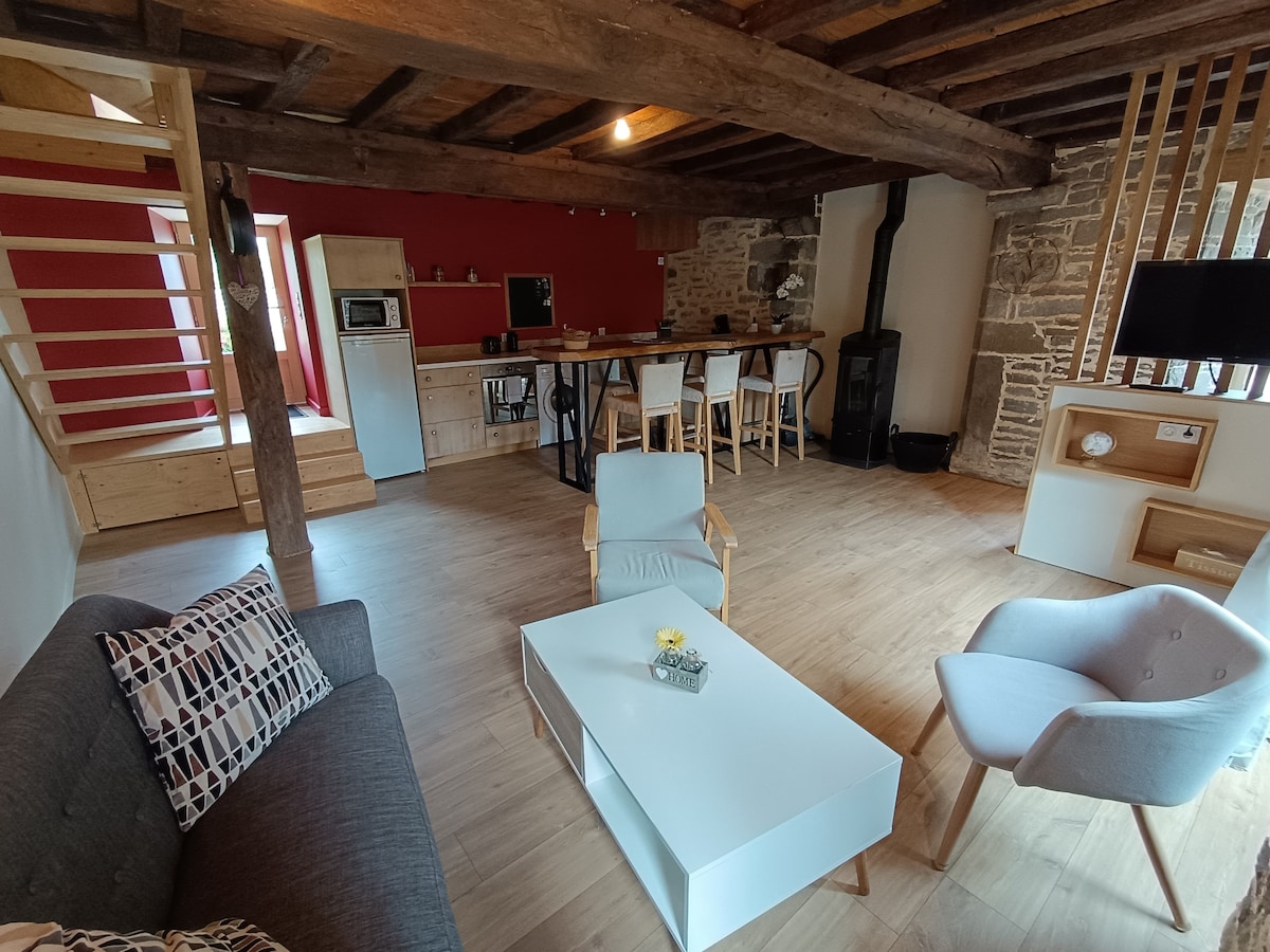 The living area features a spacious layout with wooden beams and stone walls. A grey sofa is arranged around a white coffee table, with a dining bar and high stools visible in the background. Large windows allow natural light to fill the room, enhancing the warmth of the space.