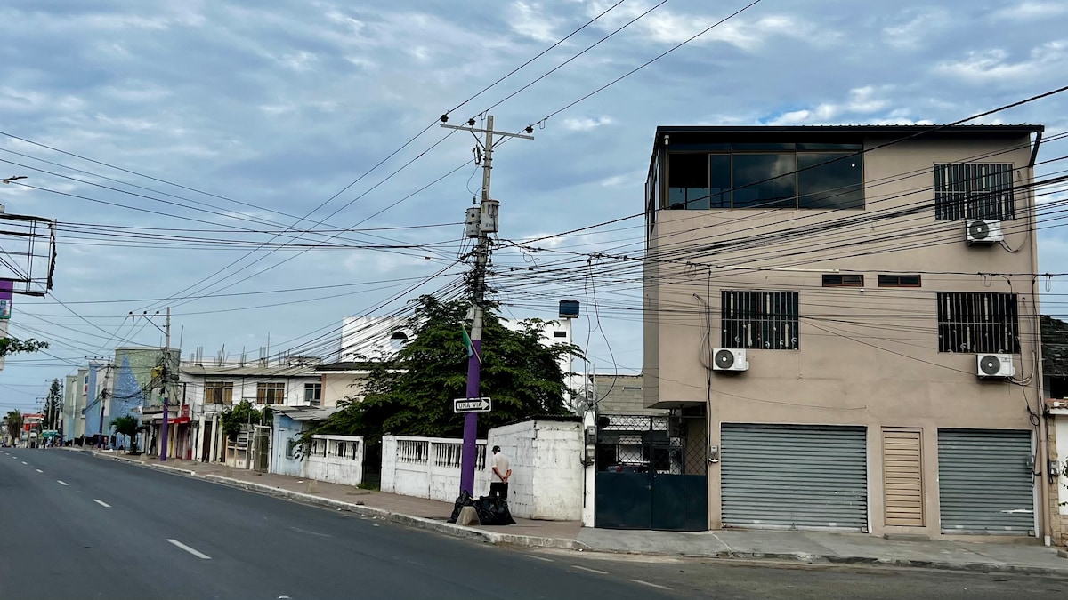 A two-story building stands along a wide street, featuring multiple air conditioning units on the exterior. Windows are positioned prominently, and power lines crisscross overhead. The adjacent street has minimal traffic, contributing to a calm atmosphere.
