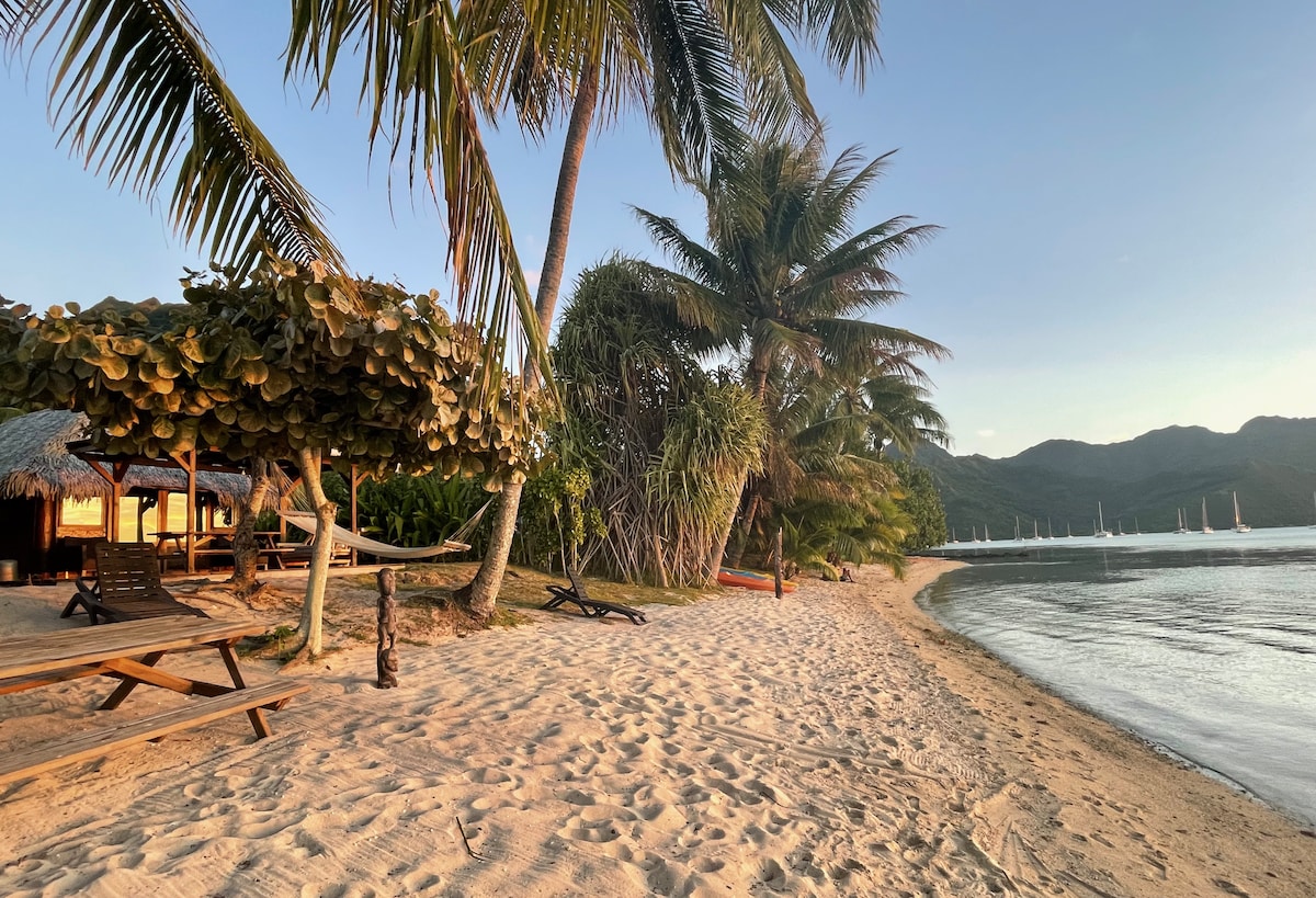 A sandy beach is lined with palm trees, providing shade and a peaceful atmosphere. Wooden loungers are positioned near a thatched-roof structure, while colorful kayaks are visible along the shore. The calm lagoon reflects the surrounding mountains in the distance.