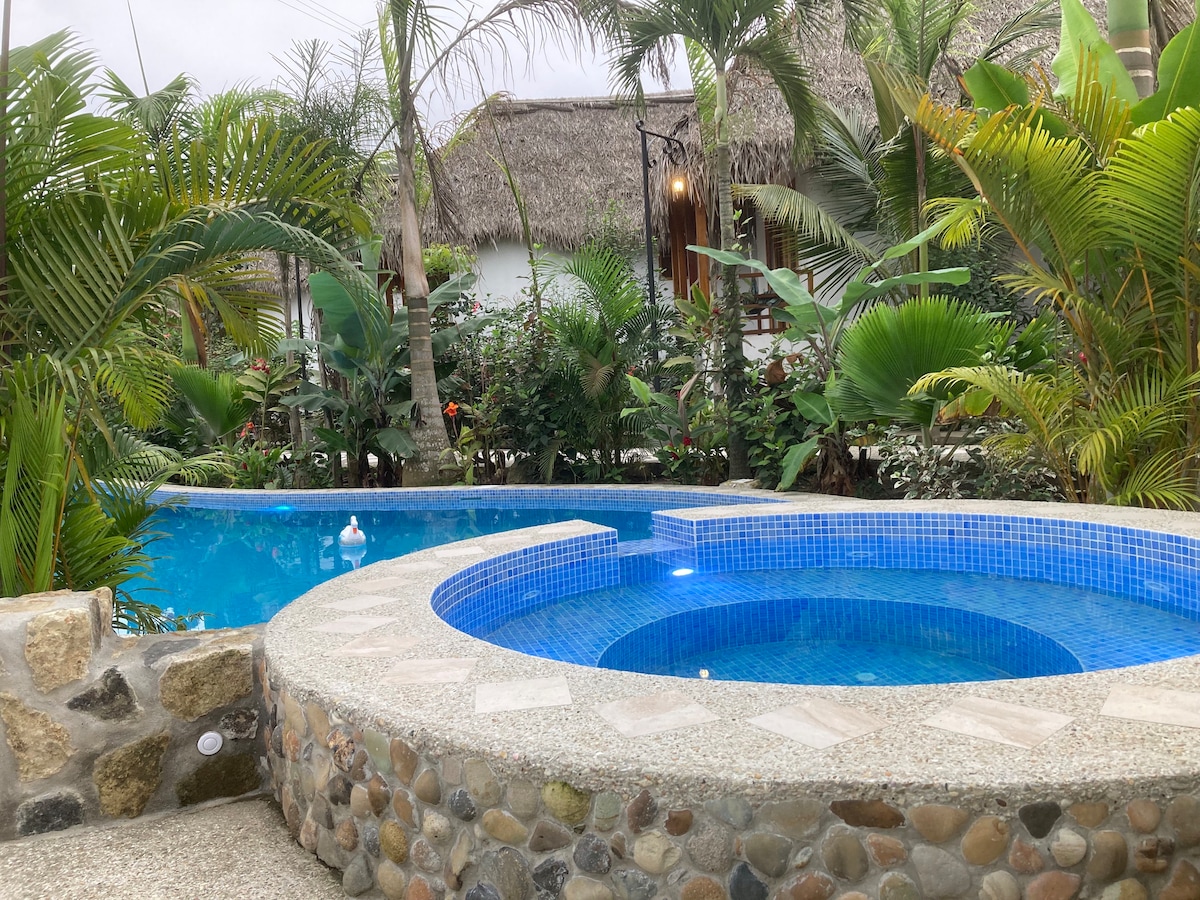 A tranquil pool area is showcased with a circular hot tub positioned next to a larger swimming pool. Lush greenery surrounds the space, including palm and tropical plants, creating a serene atmosphere. The stone and tile finishes of the pools complement the natural setting.