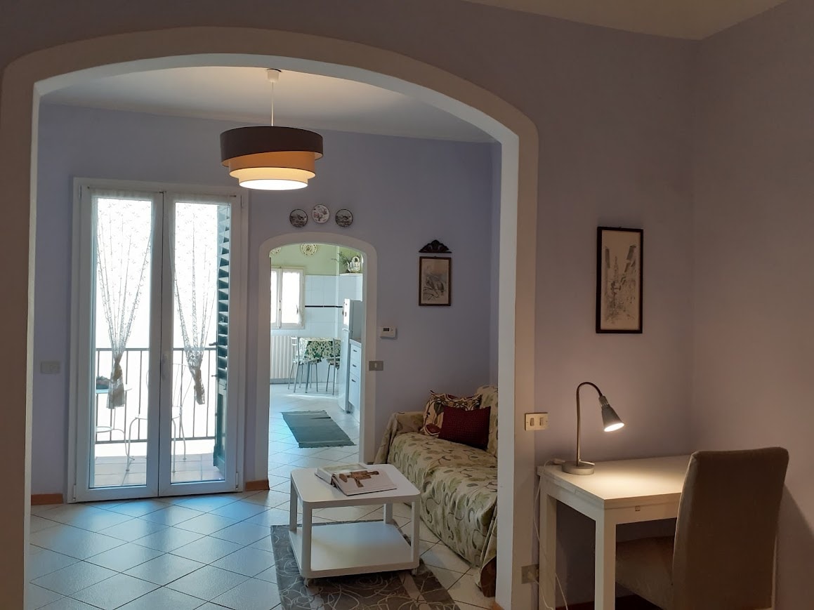 A bright living area is presented with a cozy sofa adorned with decorative cushions. A small coffee table is positioned in front of the sofa. A desk and chair are situated against the wall, with a lamp providing soft light. Large glass doors lead to a balcony.