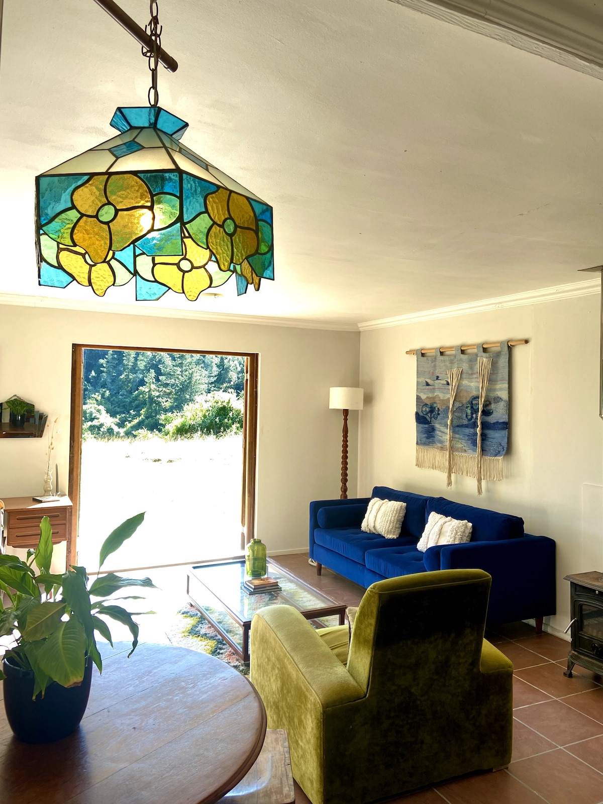A bright living area is illuminated by a colorful stained glass lamp. A blue sofa and green accent chair are positioned around a glass coffee table, with a view of the outdoor nature through the large glass door. Decorative wall hangings contribute to the cozy decor.