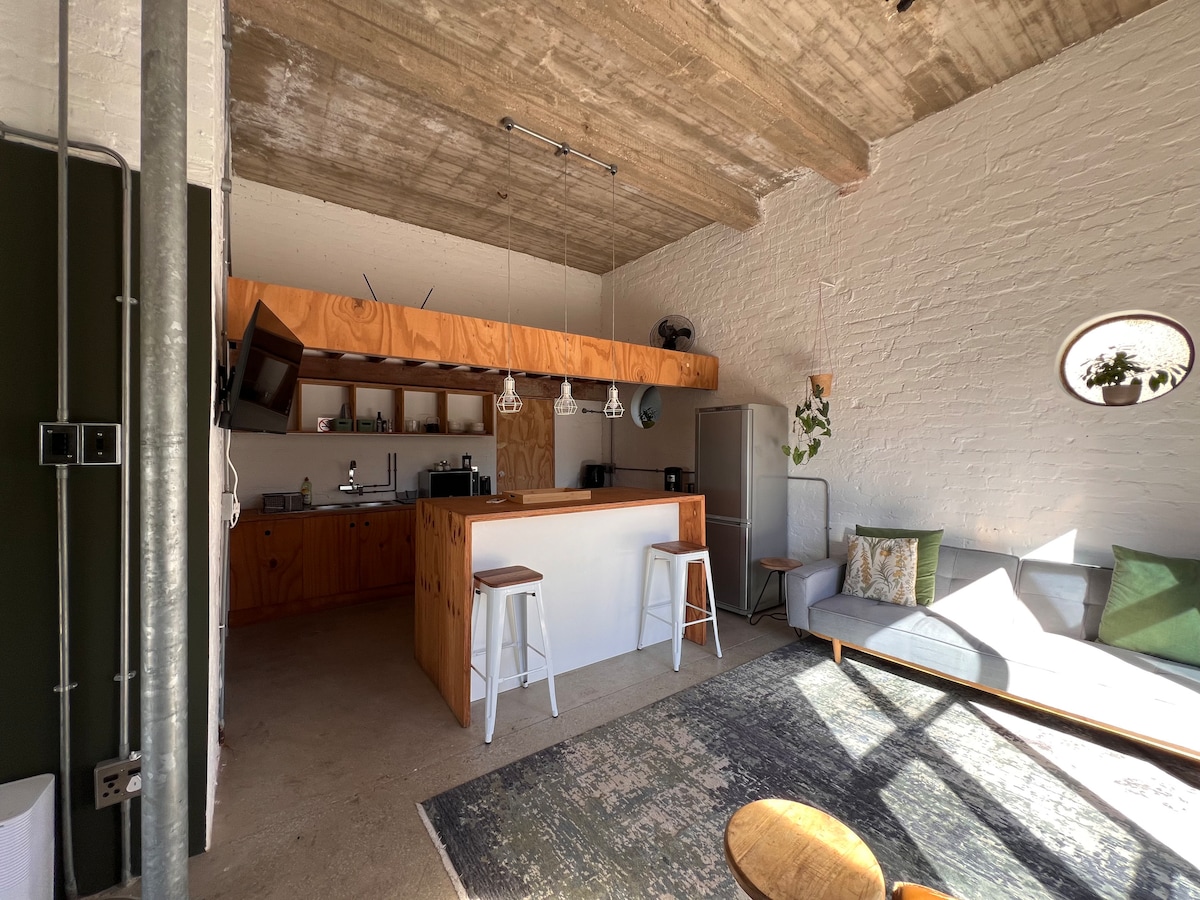 An open-plan living area features a spacious kitchen with wooden cabinetry and a breakfast bar with three stools. A cozy seating area showcases a light-toned sofa with green accent pillows. Natural light brightens the space through a circular window, highlighting the concrete ceilings and textured walls.