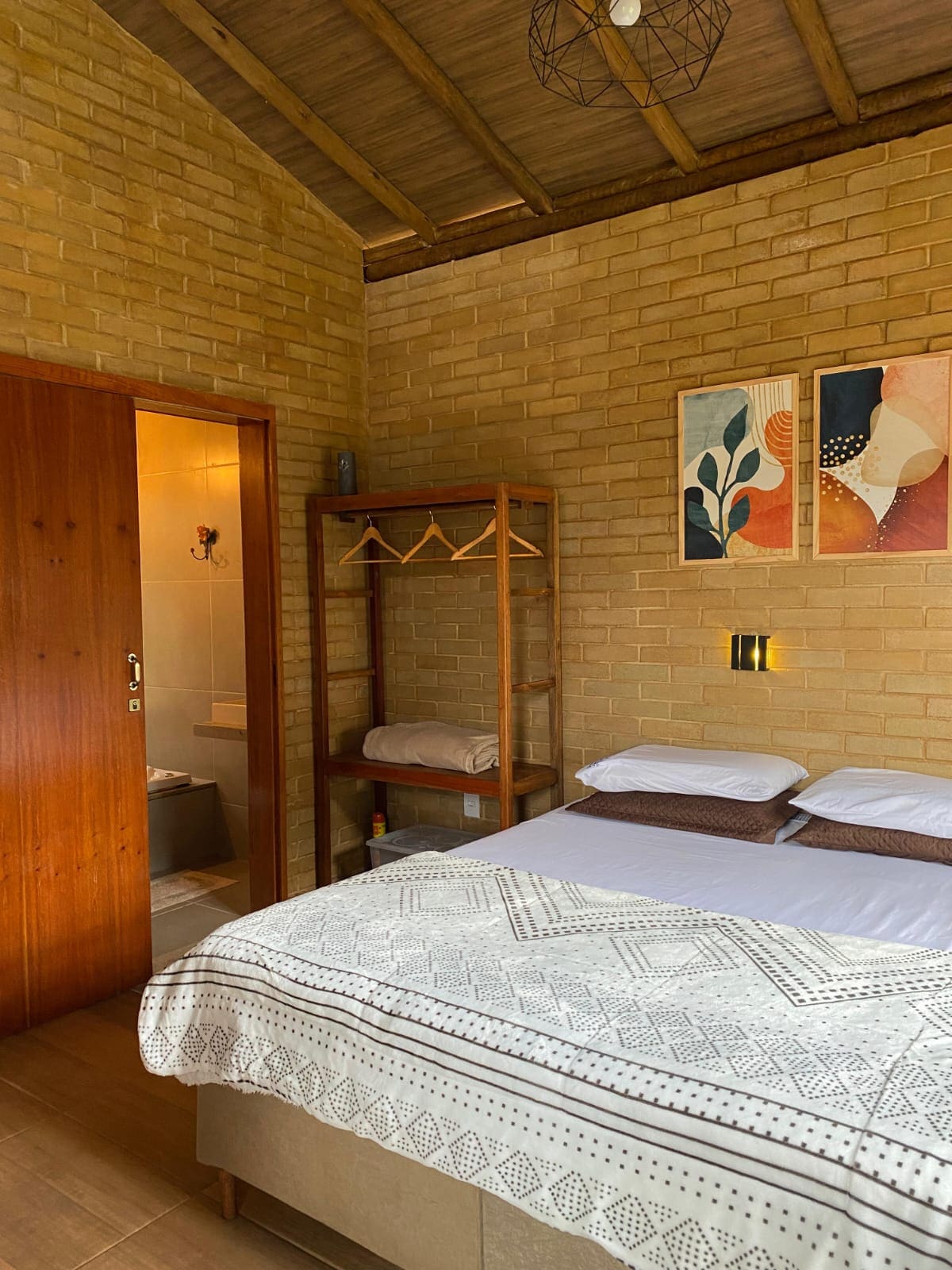 A cozy bedroom offers a spacious bed with decorative linens, complemented by earthy tones on the walls. Adjacent, a wooden rack provides space for hanging items. Artwork with abstract designs decorates the wall, adding a touch of color to the natural surroundings.