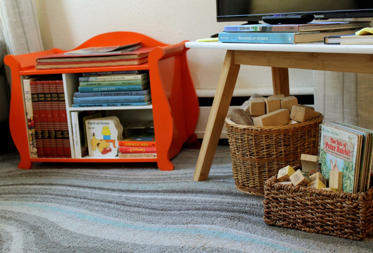 A playful corner of the living area is showcased, featuring an orange bookshelf filled with children's books. Nearby, a light wood table holds a woven basket filled with building blocks and another basket containing various storybooks, all arranged on a soft, patterned rug.