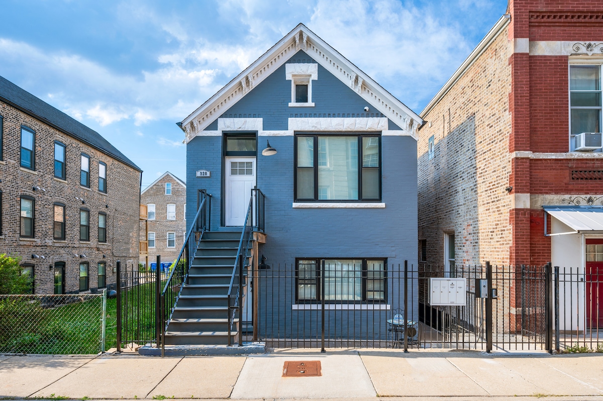 The exterior of a two-story apartment building is depicted, featuring a blue façade and white trim. A set of stairs leads to the entrance, framed by black fencing. Surrounding buildings exhibit brick and siding materials, with green space visible in the background.