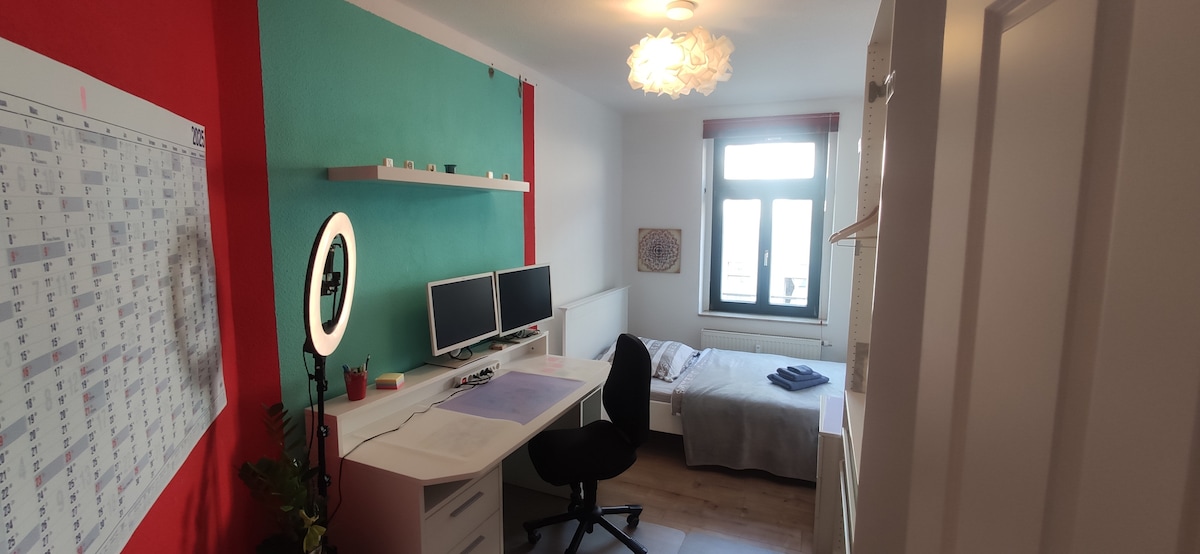 A well-organized room features a spacious bed beside a window, allowing natural light. A white desk with two monitors and a comfortable chair is in one corner, while a calendar hangs on the wall. A soft light fixture adds warmth to the space.
