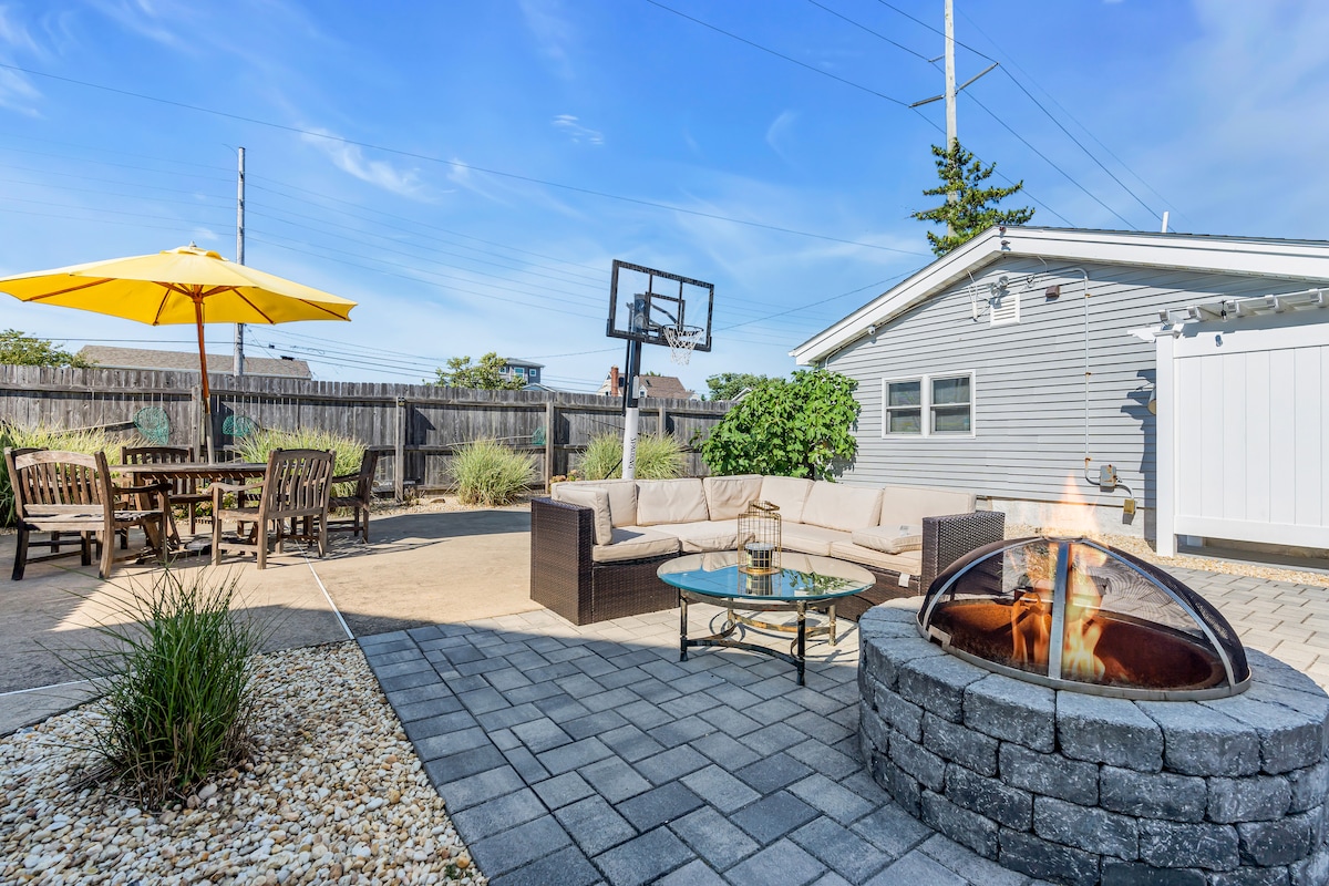 An outdoor patio area features a fire pit surrounded by stone, a glass-top coffee table, and a sectional sofa. A dining set under a yellow umbrella offers seating, while a basketball net is positioned in the background. Lush plants and decorative stones enhance the space.
