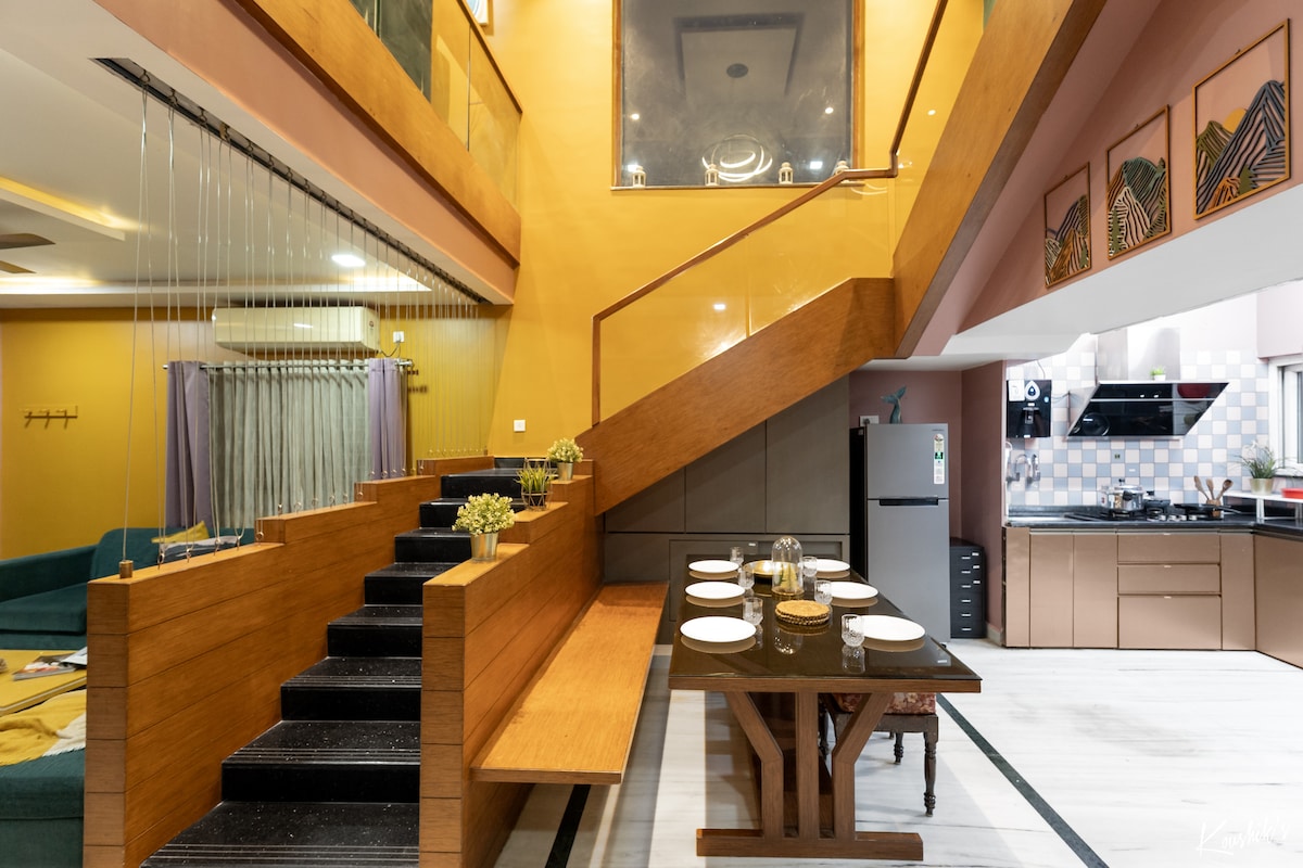 A spacious dining area features a dark wooden table set for six, complemented by elegant decor. A large staircase with wooden banisters leads to the upper level. The open layout enhances natural light, illuminating the adjacent kitchen area, which showcases modern appliances.