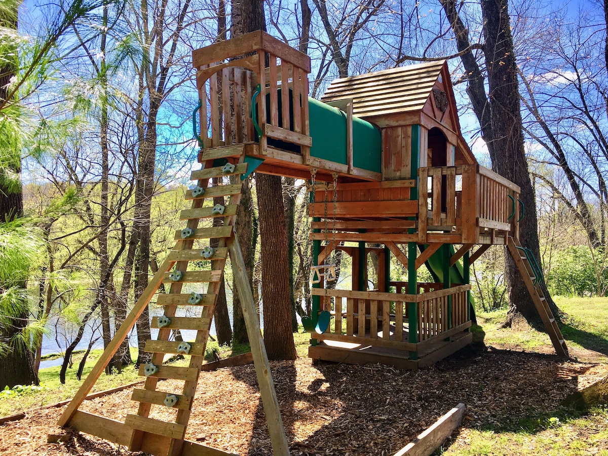 A wooden children's playground set is positioned among trees, featuring a slide, swings, and a fort-like structure. The playground is surrounded by soft mulch, with the scenic banks of the river visible in the background, offering a playful outdoor space in a natural setting.